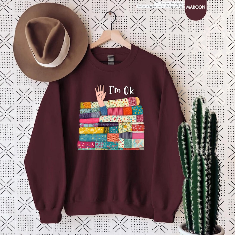 May include: Maroon sweatshirt with the text "I'm Ok" above a stack of colorful fabric. The fabric stack has a hand reaching out from it. A brown hat hangs above the sweatshirt.