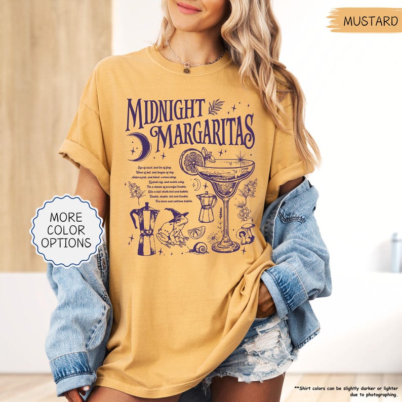 May include: Mustard-colored t-shirt with the text "Midnight Margaritas" in a dark purple font. The design includes a margarita glass, a moon, and other cocktail-related illustrations. The shirt also has the text "More Color Options".