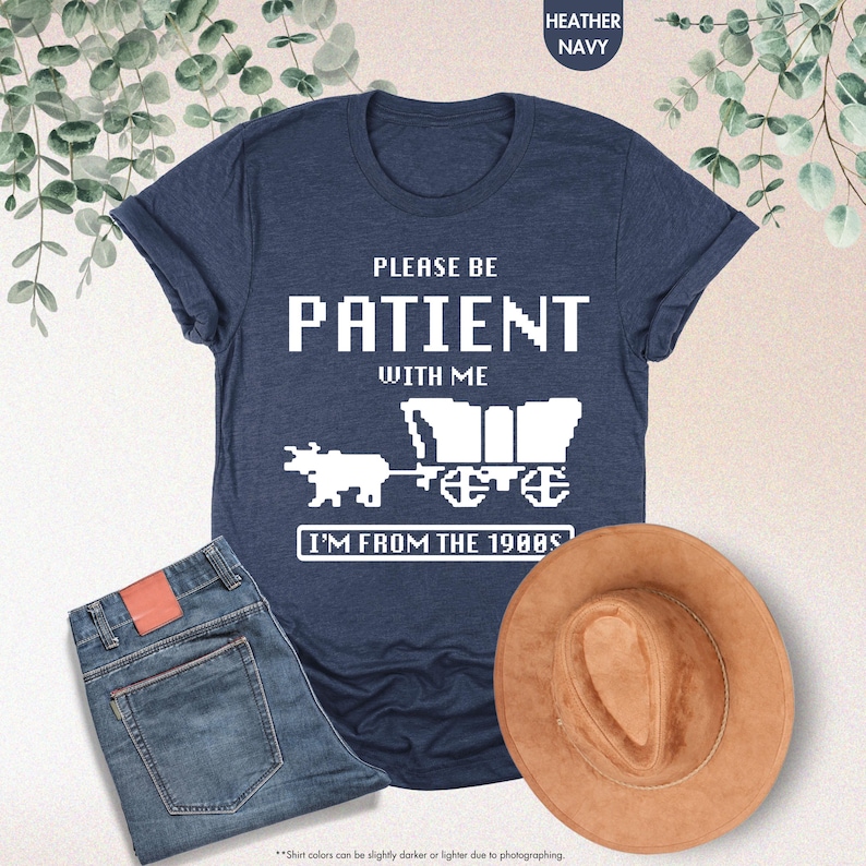 May include: Heather navy blue t-shirt with white pixelated text that reads "PLEASE BE PATIENT WITH ME, I'M FROM THE 1980S". The shirt features a pixelated wagon and oxen graphic. A pair of blue jeans and a brown hat are also in the image.