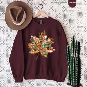 Puede incluir: Una sudadera de cuello redondo color granate con un gr&aacute;fico de hoja de arce compuesto por varios patrones oto&ntilde;ales que incluyen calabazas, girasoles y cuadros. Un sombrero fedora marr&oacute;n con una banda blanca se sit&uacute;a encima.
