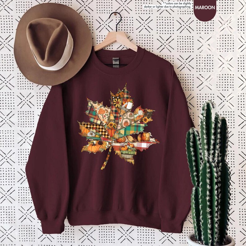 Puede incluir: Una sudadera de cuello redondo color granate con un gr&aacute;fico de hoja de arce compuesto por varios patrones oto&ntilde;ales que incluyen calabazas, girasoles y cuadros. Un sombrero fedora marr&oacute;n con una banda blanca se sit&uacute;a encima.