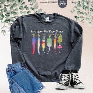 May include: A dark heather grey sweatshirt with a colorful illustration of carrots and radishes. The text "Let's Root For Each Other" is printed on the front of the sweatshirt.