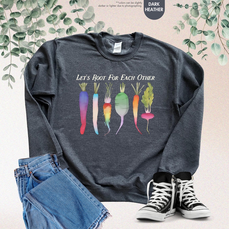 May include: A dark heather grey sweatshirt with a colorful illustration of carrots and radishes. The text "Let's Root For Each Other" is printed on the front of the sweatshirt.