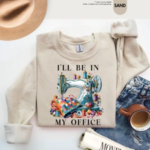 K&ouml;nnte beinhalten: Ein beiger Pullover mit dem Text "I'll be in my office" und einer bunten Illustration einer N&auml;hmaschine mit Garnrollen und Stoff.