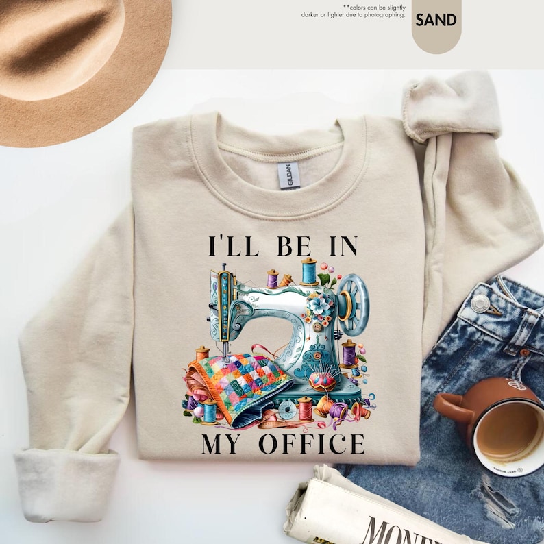 K&ouml;nnte beinhalten: Ein beiger Pullover mit dem Text "I'll be in my office" und einer bunten Illustration einer N&auml;hmaschine mit Garnrollen und Stoff.