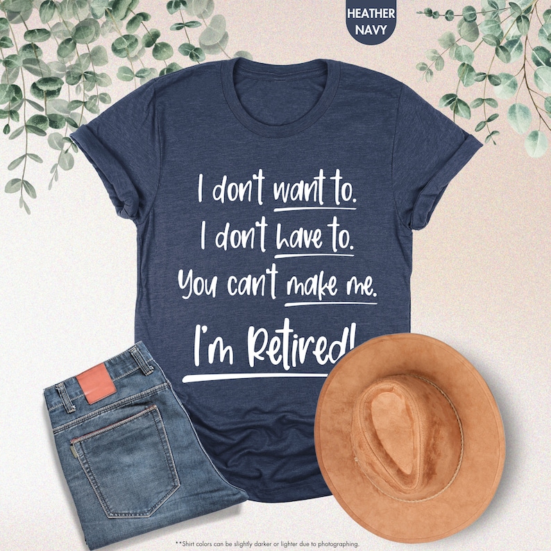 May include: Heather navy blue t-shirt featuring a retirement announcement. The text on the shirt reads: 'I don't want to. I don't have to. You can't make me. I'm Retired!'  The shirt is shown on a flat surface with a pair of blue jeans and a brown hat. The shirt is a casual, short-sleeved crew neck style.