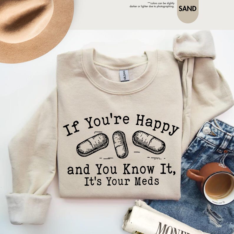 May include: A beige sweatshirt with the text "If You're Happy and You Know It, It's Your Meds" printed in black. Three black and white pill illustrations are above the text.