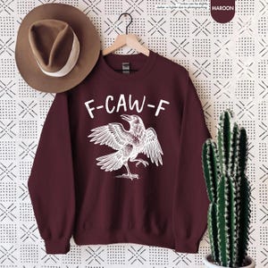 May include: Maroon sweatshirt with the text "F-CAW-F" above a white illustration of a crow with outstretched wings. A brown hat with a white band hangs to the left. A cactus in a white pot is on the right.