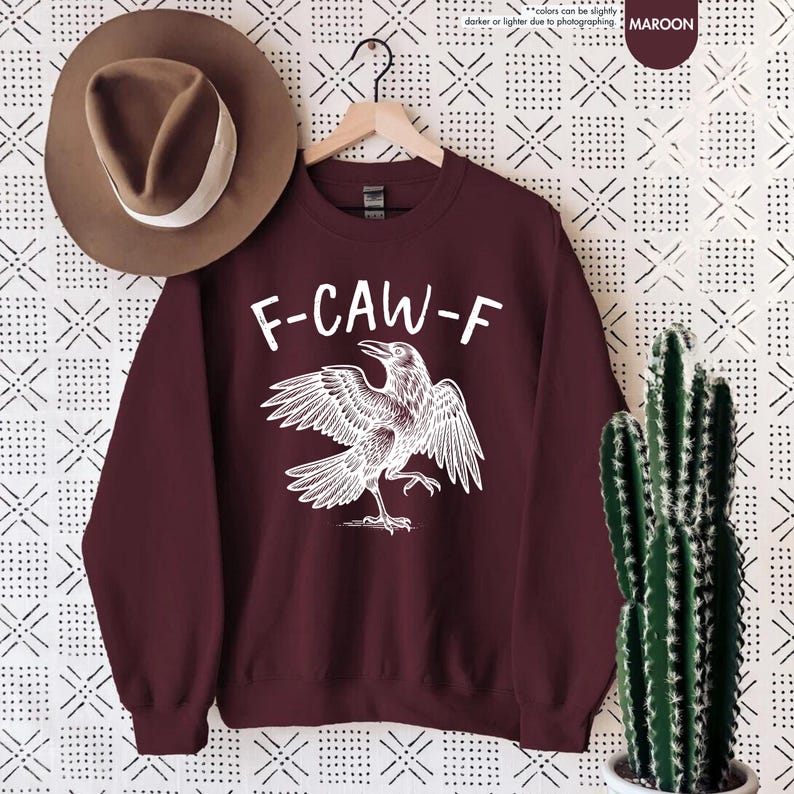 May include: Maroon sweatshirt with the text "F-CAW-F" above a white illustration of a crow with outstretched wings. A brown hat with a white band hangs to the left. A cactus in a white pot is on the right.