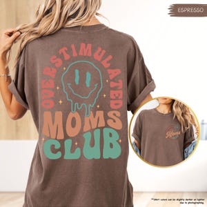 K&ouml;nnte beinhalten: Espressofarbenes T-Shirt mit dem Schriftzug "OVERSTIMULATED MOMS CLUB" in einem Retro-Schriftstil. Das Design umfasst ein schmelzendes Smiley-Gesicht und Sternakzente. Das Shirt hat hochgekrempelte &Auml;rmel und eine lockere Passform.