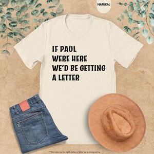 May include: Cream-colored t-shirt with the text "IF PAUL WERE HERE WE'D BE GETTING A LETTER" in black. The shirt is paired with blue jeans and a brown hat. The background has a natural, neutral tone.