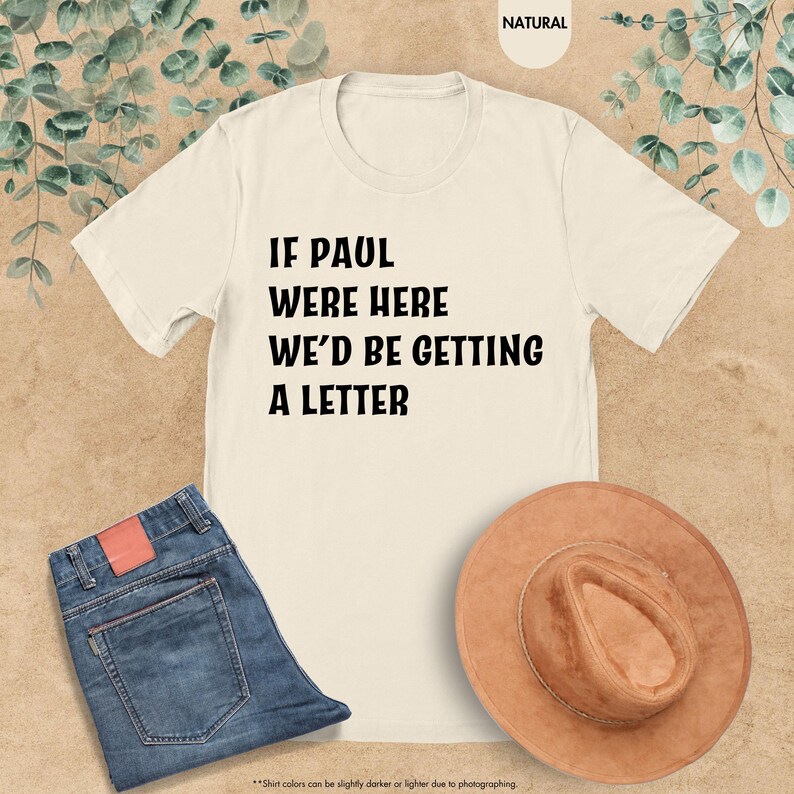 May include: Cream-colored t-shirt with the text "IF PAUL WERE HERE WE'D BE GETTING A LETTER" in black. The shirt is paired with blue jeans and a brown hat. The background has a natural, neutral tone.