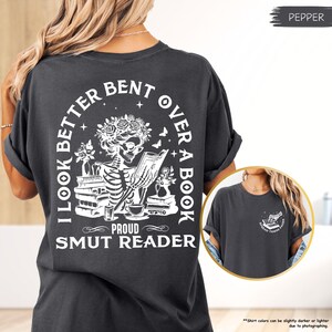 以下が含まれることがあります： 本を読んでいる骸骨の白いグラフィックが描かれた黒いTシャツ。シャツのテキストは「I look better bent over a book, Proud Smut Reader」です。
