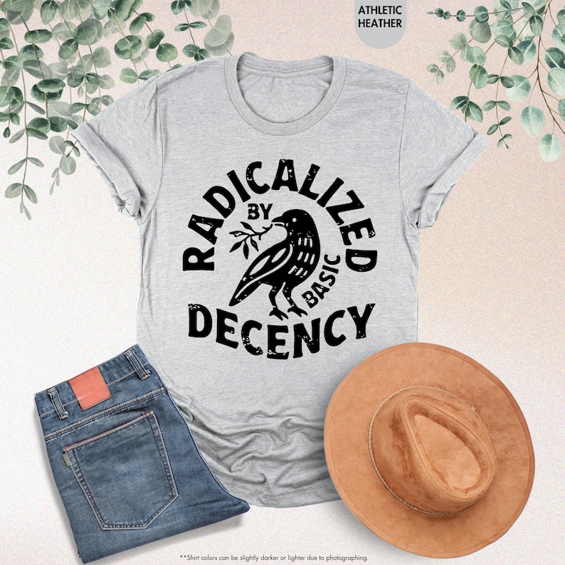 May include: Athletic heather gray t-shirt with the text "RADICALIZED BY BASIC DECENCY" in a circular design around a raven holding a sprig. The image also shows blue jeans and a brown hat.