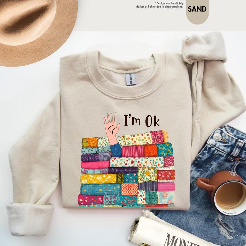 May include: Sand-colored sweatshirt with the text "I'm Ok" and a graphic of a hand emerging from a stack of colorful books. The books have various patterns and colors. The sweatshirt is paired with blue jeans and a brown mug.