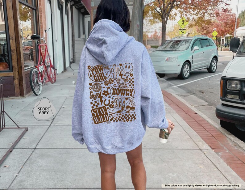 K&ouml;nnte beinhalten: Ein hellgrauer Kapuzenpullover mit einem braunen Grafikdesign auf dem R&uuml;cken. Das Design enth&auml;lt die W&ouml;rter "Cow Girl", "Howdy" und "Stay Wild" sowie verschiedene Illustrationen. Der Kapuzenpullover ist mit "Sport Grey" gekennzeichnet.