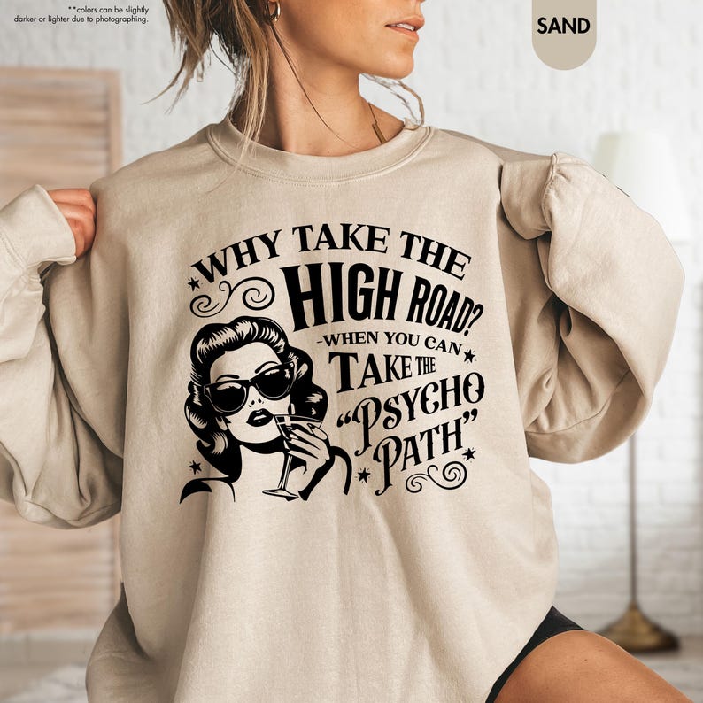 May include: Sand-colored sweatshirt with the text "WHY TAKE THE HIGH ROAD? WHEN YOU CAN TAKE THE PSYCHO PATH" and a black and white graphic of a woman wearing sunglasses and holding a drink.