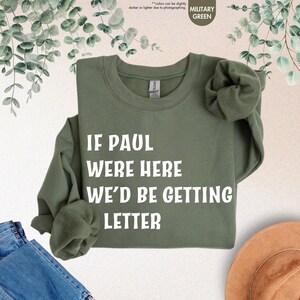 May include: Military green sweatshirt with the text "IF PAUL WERE HERE WE'D BE GETTING LETTER" in white block letters. The long-sleeved crewneck shirt is folded, with a hint of denim and a hat in the background.