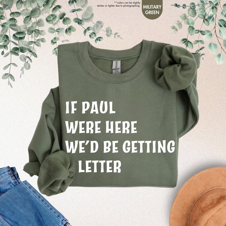 May include: Military green sweatshirt with the text "IF PAUL WERE HERE WE'D BE GETTING LETTER" in white block letters. The long-sleeved crewneck shirt is folded, with a hint of denim and a hat in the background.