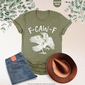 May include: Heather olive green t-shirt with the text "F-CAW-F" and a white raven illustration. The image also includes blue jeans and a brown hat. The shirt's color may vary slightly due to photography.