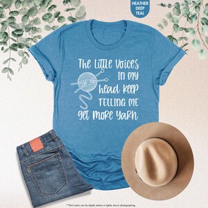May include: A blue t-shirt with a white graphic that says "The Little Voices In My Head Keep Telling Me Get More Yarn". The graphic also includes a drawing of a ball of yarn and knitting needles.