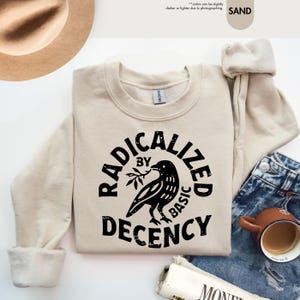 May include: Sand-colored sweatshirt with the text "RADICALIZED BY BASIC DECENCY" in a black, circular design featuring a raven holding a sprig. The sweatshirt is paired with blue jeans, a brown hat, and a coffee mug.