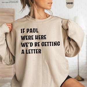 May include: Sand-colored sweatshirt with the text "IF PAUL WERE HERE WE'D BE GETTING A LETTER" in black. The long-sleeved crew neck top is made of a soft material. The sweatshirt is a casual style.