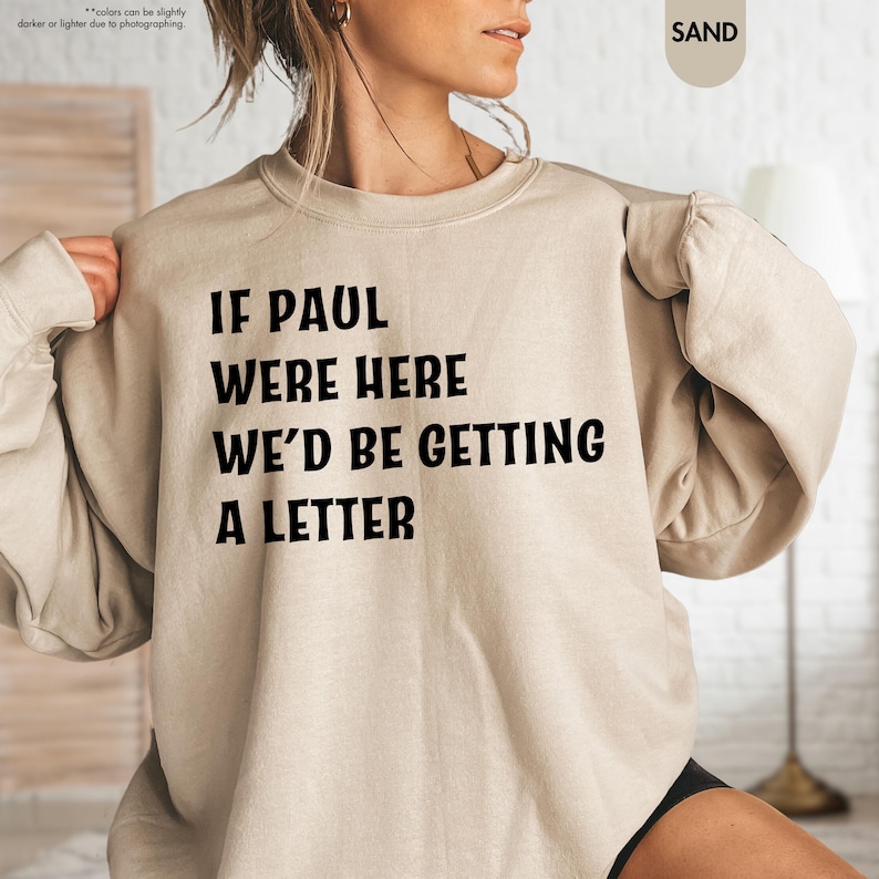 May include: Sand-colored sweatshirt with the text "IF PAUL WERE HERE WE'D BE GETTING A LETTER" in black. The long-sleeved crew neck top is made of a soft material. The sweatshirt is a casual style.