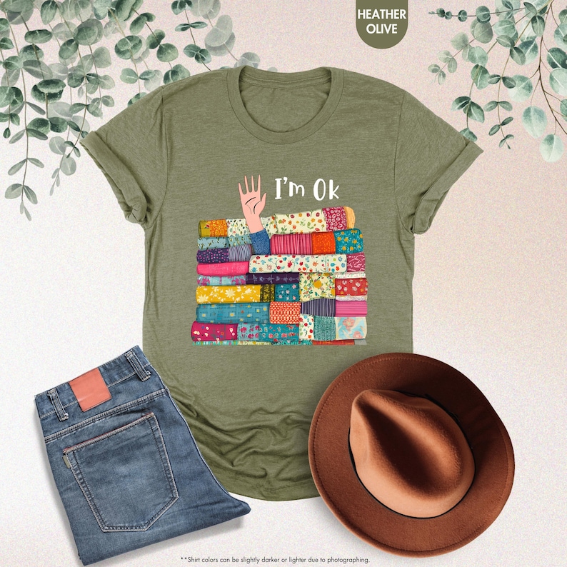 May include: Heather olive green t-shirt with the text "I'm Ok" and a graphic of a hand and a stack of colorful patchwork quilts. The image also includes blue jeans and a brown hat.
