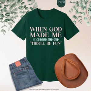 Puede incluir: Una camiseta verde oscuro con el texto "WHEN GOD MADE ME HE GRINNED AND SAID 'THIS'LL BE FUN'" en letras rosas. La imagen tambi&eacute;n incluye unos vaqueros azules y un sombrero de vaquero marr&oacute;n.