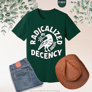 May include: A dark green t-shirt with the words "RADICALIZED BY BASIC DECENCY" in a circular design around a dove graphic. The image also includes blue jeans and a brown cowboy hat.