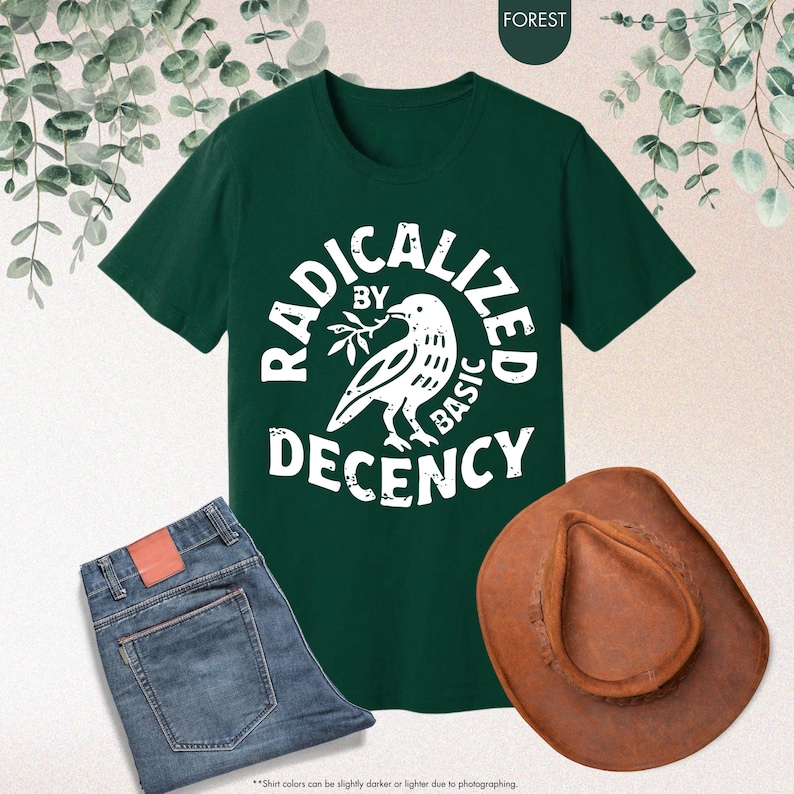 May include: A dark green t-shirt with the words "RADICALIZED BY BASIC DECENCY" in a circular design around a dove graphic. The image also includes blue jeans and a brown cowboy hat.