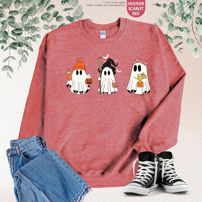 May include: A heather scarlet crewneck sweatshirt featuring a graphic of three white ghosts. The first ghost wears an orange beanie and holds a drink and pumpkin bucket. The second wears a witch hat and holds a broom. The third wears a pink headband.