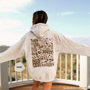 K&ouml;nnte beinhalten: Ein sandfarbener Kapuzenpullover mit einem braunen Grafikdesign auf dem R&uuml;cken. Das Design enth&auml;lt die W&ouml;rter "COW GIRL", "HOWDY" und "STAY WILD" sowie Bilder von einem Kaktus, Stiefeln und einem Smiley. Der Hoodie hat eine Kapuze und lange &Auml;rmel.