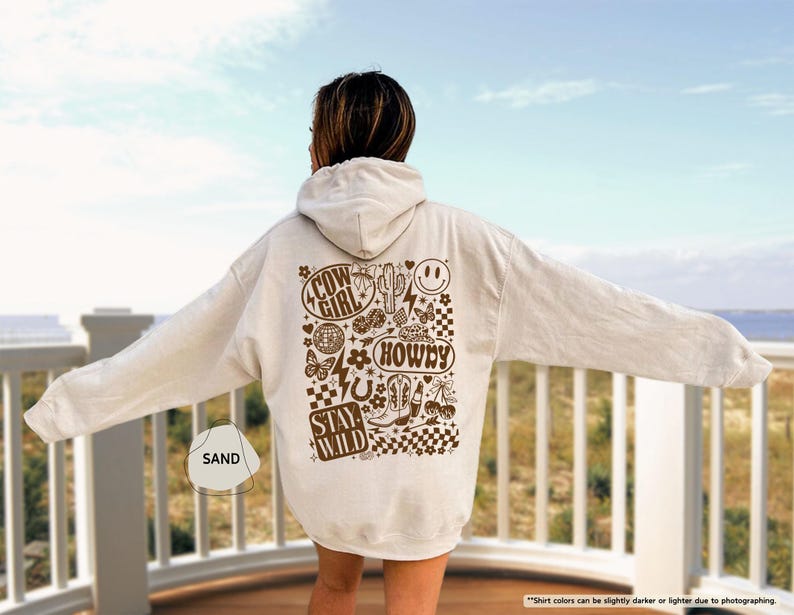 K&ouml;nnte beinhalten: Ein sandfarbener Kapuzenpullover mit einem braunen Grafikdesign auf dem R&uuml;cken. Das Design enth&auml;lt die W&ouml;rter "COW GIRL", "HOWDY" und "STAY WILD" sowie Bilder von einem Kaktus, Stiefeln und einem Smiley. Der Hoodie hat eine Kapuze und lange &Auml;rmel.