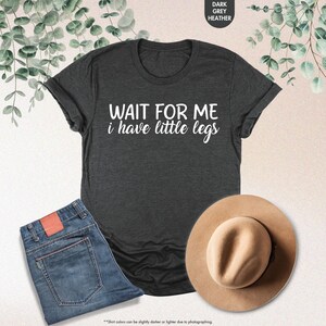 Pode incluir: T-shirt cinza escuro com a frase branca "WAIT FOR ME i have little legs". A imagem tamb&eacute;m inclui jeans azuis e um chap&eacute;u bege. &Eacute; uma pe&ccedil;a de roupa casual.