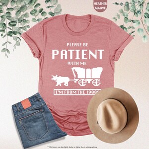 May include: Heather mauve t-shirt with the text "PLEASE BE PATIENT WITH ME" and a pixelated wagon graphic. The shirt is paired with blue jeans and a tan fedora hat. A fun, retro-themed outfit.