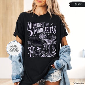 May include: Black t-shirt with a white graphic design that says "Midnight Margaritas". The design includes a cocktail glass, moon, and other illustrations. The shirt also has the text "More Color Options" on a white oval.