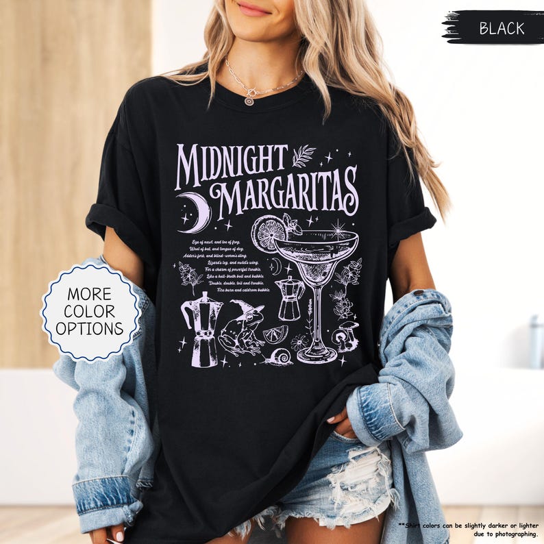 May include: Black t-shirt with a white graphic design that says "Midnight Margaritas". The design includes a cocktail glass, moon, and other illustrations. The shirt also has the text "More Color Options" on a white oval.