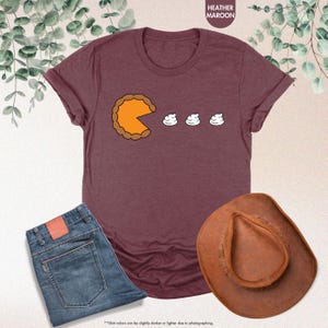 May include: A heather maroon t-shirt with a graphic of a pumpkin pie with a bite taken out of it and three dollops of whipped cream. The text "Shirt colors can be slightly darker or lighter due to photographing." is at the bottom of the image.