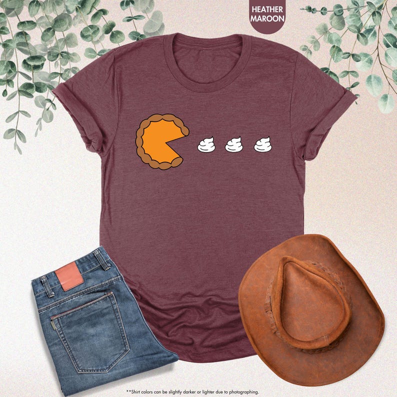 May include: A heather maroon t-shirt with a graphic of a pumpkin pie with a bite taken out of it and three dollops of whipped cream. The text "Shirt colors can be slightly darker or lighter due to photographing." is at the bottom of the image.