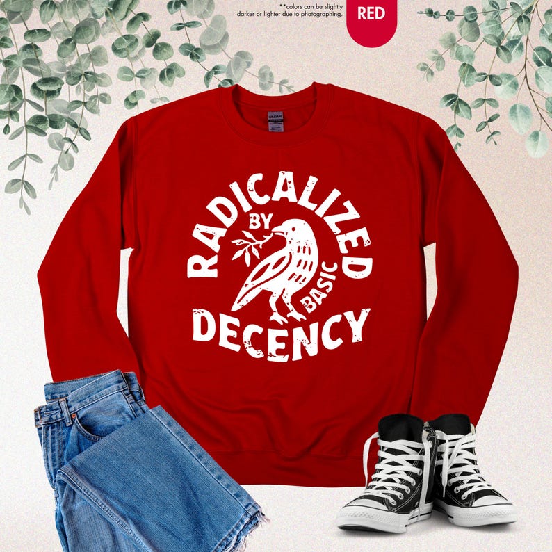May include: A red sweatshirt with the text "RADICALIZED BY BASIC DECENCY" in a white circular design featuring a dove holding an olive branch. The image also shows blue jeans and black canvas sneakers.
