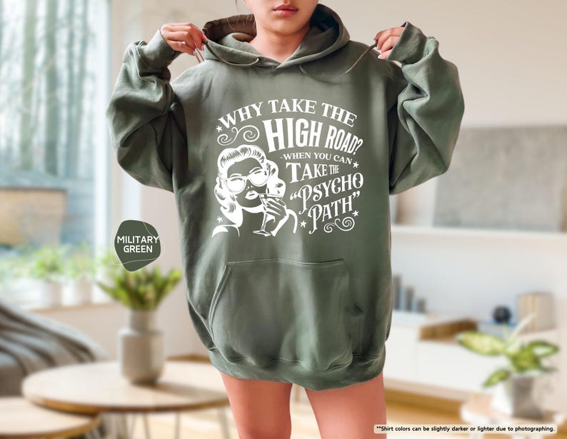 May include: Military green hoodie with a front pocket and a drawstring hood. The white graphic on the front reads "WHY TAKE THE HIGH ROAD? WHEN YOU CAN TAKE THE PSYCHO PATH" with a vintage-style illustration of a woman.