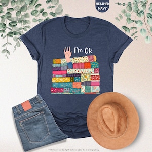 May include: Heather navy blue t-shirt with the text "I'm Ok" and a graphic of a hand and a stack of colorful fabric. The image also includes a pair of blue jeans and a brown hat.
