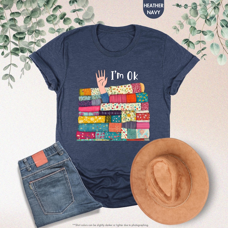 May include: Heather navy blue t-shirt with the text "I'm Ok" and a graphic of a hand and a stack of colorful fabric. The image also includes a pair of blue jeans and a brown hat.