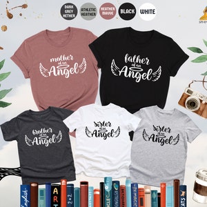 Op de afbeelding: Vijf T-shirts in verschillende kleuren en ontwerpen. De shirts hebben de tekst "moeder van een engel", "vader van een engel", "broer van een engel" en "zus van een engel" erop gedrukt. De shirts zijn in een rij gerangschikt, met het shirt "moeder van een engel" vooraan.