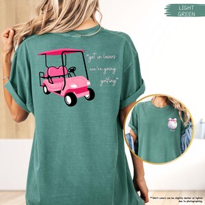 May include: A light green t-shirt with a pink golf cart graphic and the text "get in losers we're going golfing". The shirt also features a small graphic of a golf ball with a pink bow.