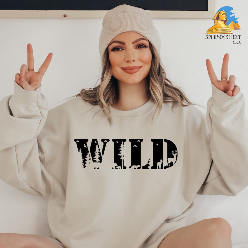 May include: A beige sweatshirt with the word "WILD" printed in black. The letters are filled with images of trees, animals, and birds.