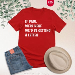 May include: A red t-shirt with the text "IF PAUL WERE HERE WE'D BE GETTING A LETTER" in white. The shirt is paired with blue jeans and a beige hat. The word "RED" is in a red circle in the top right corner.