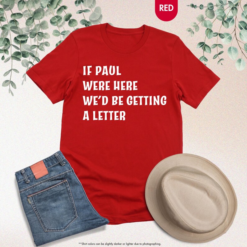 May include: A red t-shirt with the text "IF PAUL WERE HERE WE'D BE GETTING A LETTER" in white. The shirt is paired with blue jeans and a beige hat. The word "RED" is in a red circle in the top right corner.